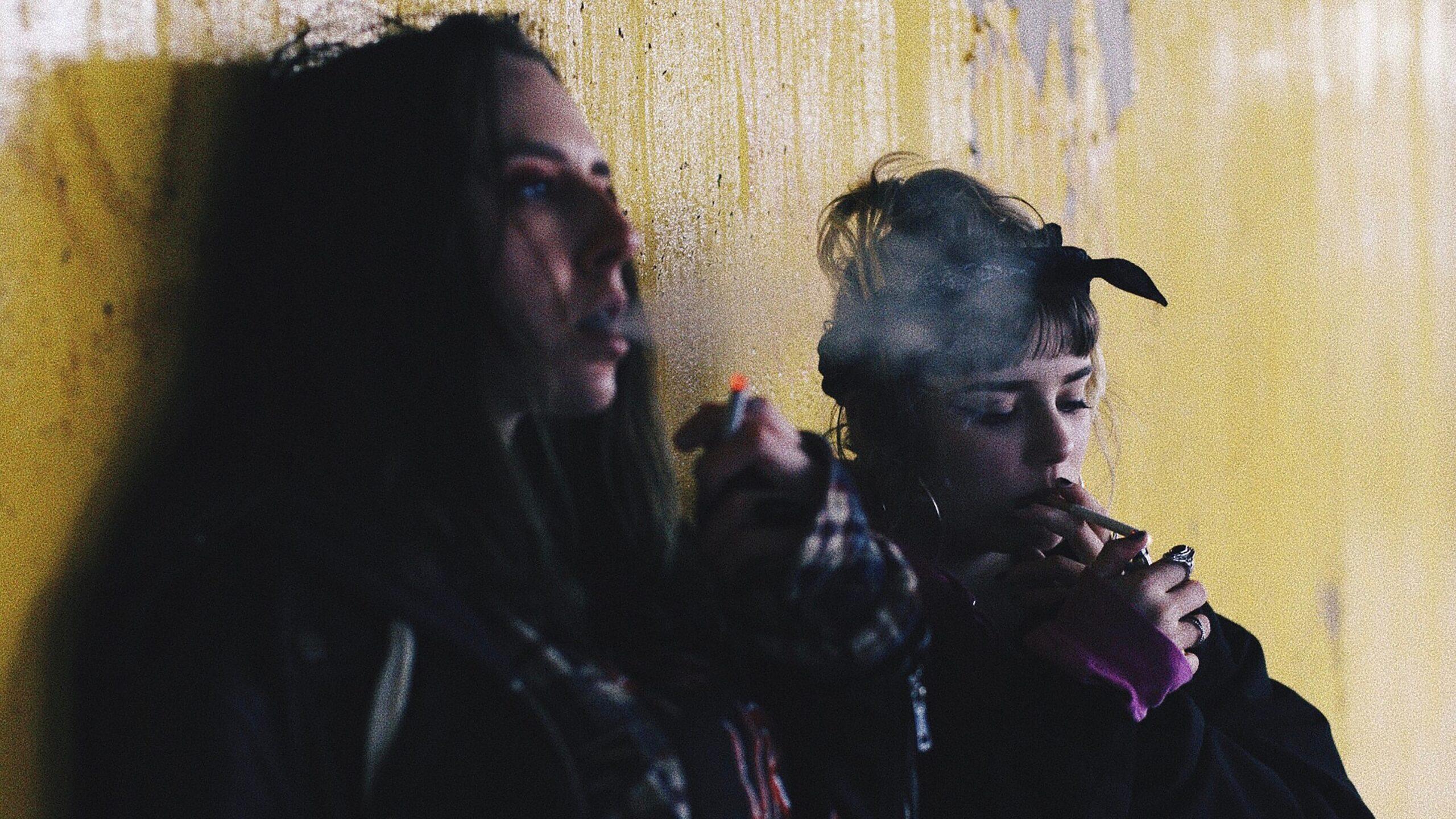 two women smoking against a yellow wall